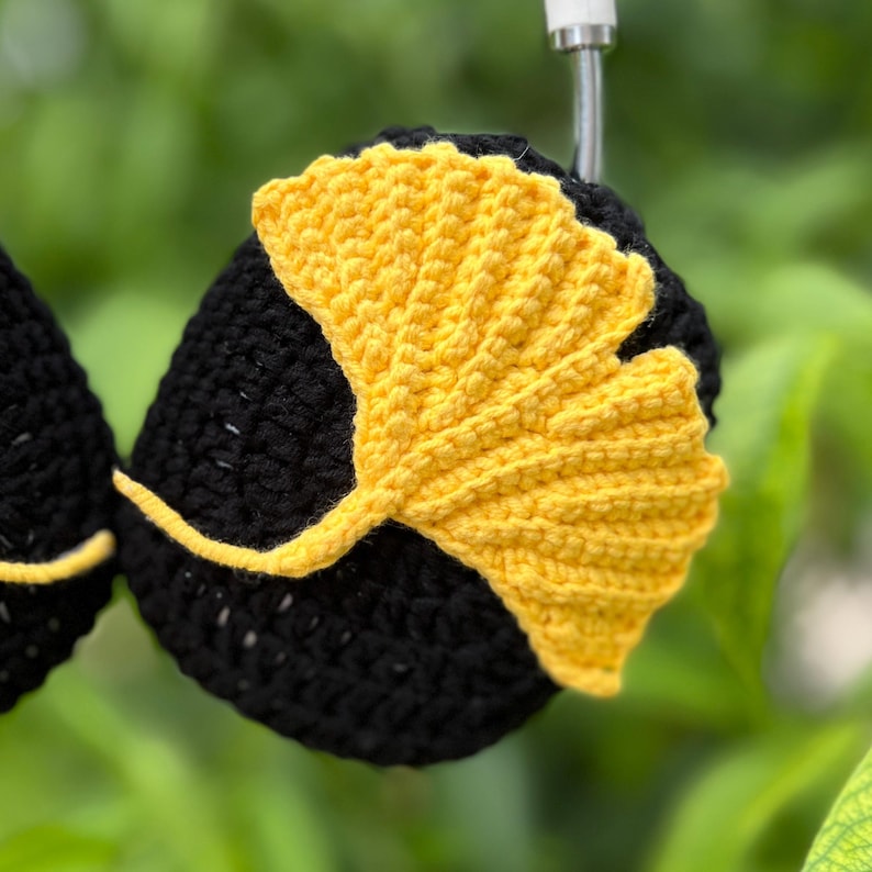 Pu&ograve; includere: Cuffie nere all'uncinetto con un design a foglia di ginkgo giallo brillante. La foglia &egrave; realizzata all'uncinetto in modo intricato, aggiungendo un tocco unico e fatto a mano. Le cuffie sono ambientate su uno sfondo verde sfocato, evidenziando i colori e le trame contrastanti.