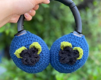 May include: Blue crochet headphone covers with a black and green floral design. The headphones are black with the brand name "SONY" visible on the earcups. The background is a blurred green.