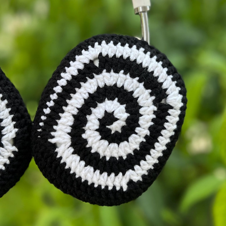 May include: Black and white crochet headphone covers. The covers feature a circular pattern of alternating black and white stripes. The headphones have a silver metal band.