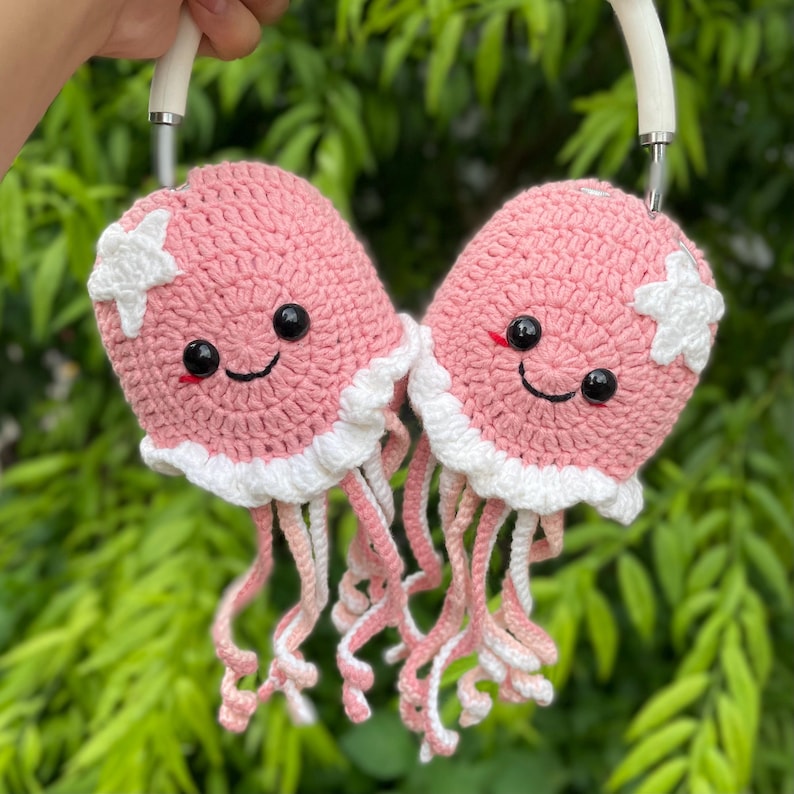 Puede incluir: Un par de fundas de crochet rosas para auriculares con forma de medusas sonrientes, con volantes blancos y detalles de estrellas. Las medusas tienen ojos negros y tent&aacute;culos rosas y blancos. Los auriculares se sujetan con una diadema blanca.