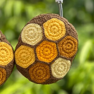 Puede incluir: Una funda de auriculares de ganchillo marr&oacute;n y amarillo, con un dise&ntilde;o de panal. La funda presenta una serie de hex&aacute;gonos amarillos y crema, rodeados por un borde marr&oacute;n. El dise&ntilde;o recuerda a un panal de abejas.