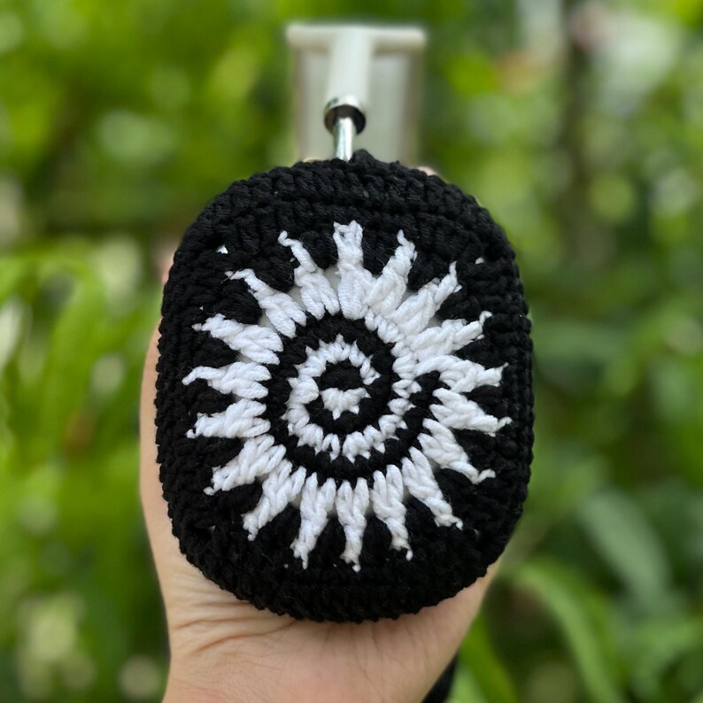 May include: Black and white crochet headphone case featuring a spiral sun design. The case is made of a soft, textured yarn and has a loop for hanging. The design is a bold contrast against the black background.