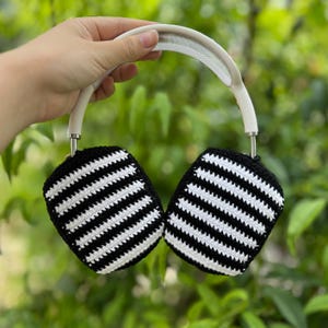 May include: White and black striped crochet headphone covers. The covers are attached to a white headband with a mesh top. The ear covers are black with white horizontal stripes. The headphones are held up against a green leafy background.