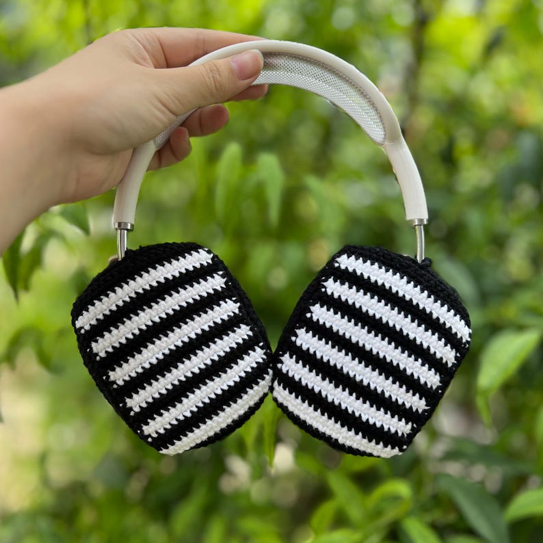 May include: White and black striped crochet headphone covers. The covers are attached to a white headband with a mesh top. The ear covers are black with white horizontal stripes. The headphones are held up against a green leafy background.