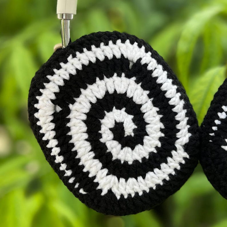 May include: Black and white crocheted headphone covers with a spiral design. The covers are circular and appear to be handmade, with a repeating pattern of black and white concentric circles. The background is a blurred green.