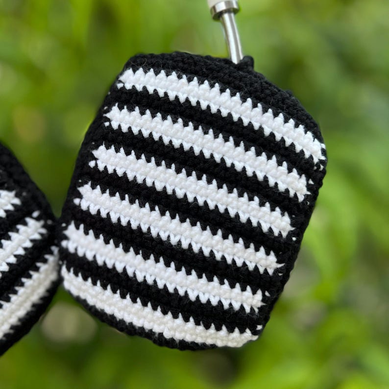 May include: Close-up of a black and white striped crochet item. The item is a small, rounded shape with a black border and a silver metal attachment. The crochet pattern features alternating black and white horizontal stripes.