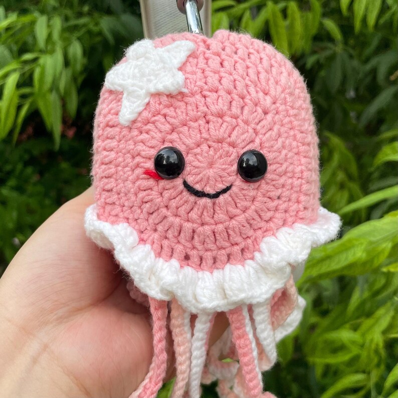 Puede incluir: Un llavero de medusa rosa tejido a mano con una estrella blanca y una cara sonriente. La medusa tiene ojos negros, boca roja y tent&aacute;culos blancos y rosas. El llavero est&aacute; sujeto a un cierre de metal plateado.