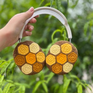 Puede incluir: Auriculares de crochet con dise&ntilde;o de panal de abeja. Los auriculares son marrones con patrones hexagonales amarillos y dorados. La diadema es blanca. Los auriculares se sostienen contra un fondo verde borroso de follaje.