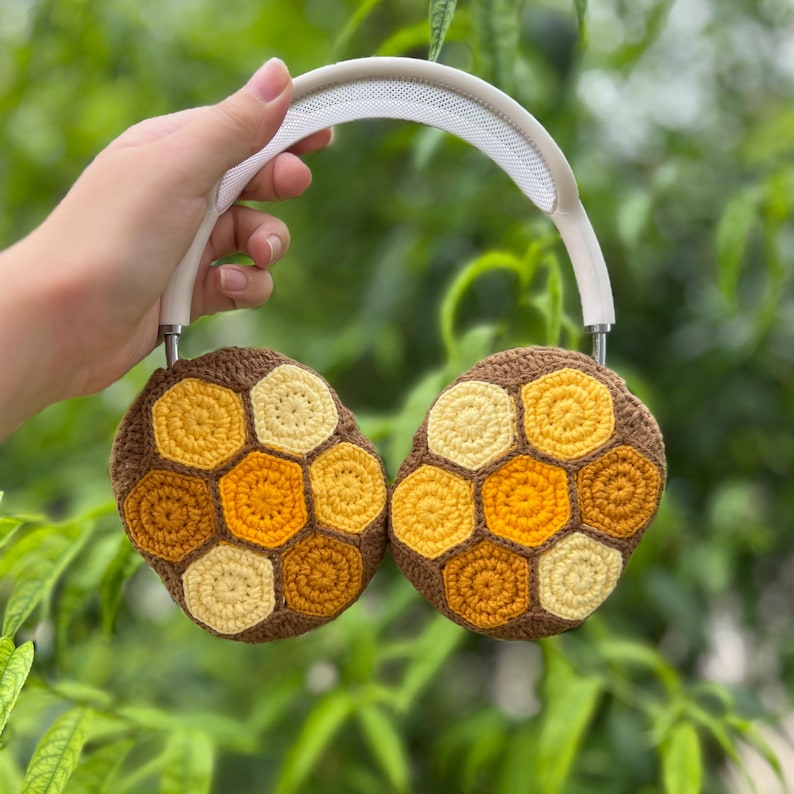 Puede incluir: Auriculares de crochet con dise&ntilde;o de panal de abeja. Los auriculares son marrones con patrones hexagonales amarillos y dorados. La diadema es blanca. Los auriculares se sostienen contra un fondo verde borroso de follaje.
