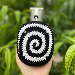 May include: A black and white crocheted bottle stopper cover with a spiral design. The cover is held in a hand against a blurred green background. The stopper has a silver metal stem.