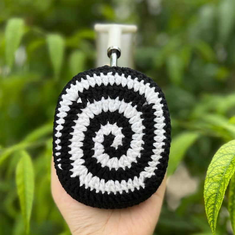 May include: A black and white crocheted bottle stopper cover with a spiral design. The cover is held in a hand against a blurred green background. The stopper has a silver metal stem.