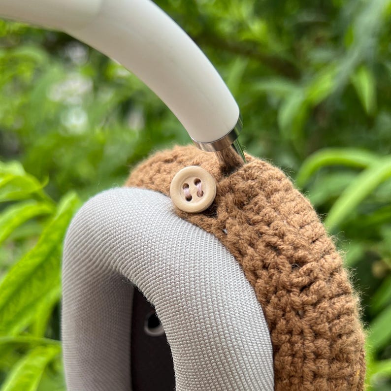 Puede incluir: Primer plano de una funda de auriculares marr&oacute;n tejida a ganchillo con un bot&oacute;n beige. La funda est&aacute; sujeta a una diadema blanca. Los auriculares tienen una almohadilla gris. El fondo es follaje verde borroso.