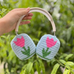 May include: Crocheted headphones with light blue ear covers featuring a pink tulip design with green leaves and stems. The headband is white. The headphones are held against a blurred green background.