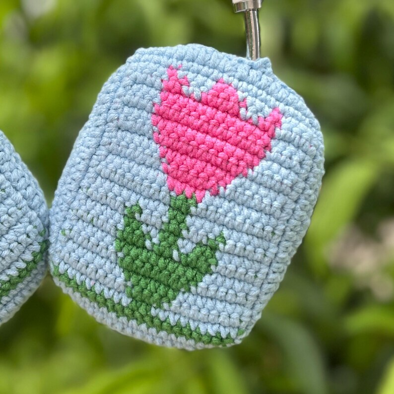 May include: A light blue crocheted case featuring a pink flower design with green leaves and stem. The case is made of soft yarn and has a loop at the top. The background is blurred green foliage.