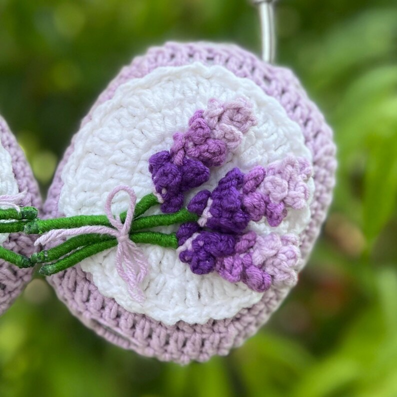 May include: Crocheted lavender-themed item featuring a white center, a lavender-colored border, and a detailed lavender flower design in shades of purple. The flower stems are green, tied with a light purple bow. The item is handmade.