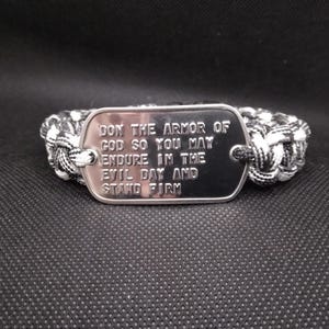 May include: A black and white braided bracelet with a silver tag. The tag has the engraved text: "DON THE ARMOR OF GOD SO YOU MAY ENDURE IN THE EVIL DAY AND STAND FIRM."
