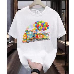 K&ouml;nnte beinhalten: Ein wei&szlig;es kurz&auml;rmeliges T-Shirt mit einem bunten Grafikdruck eines Cartoon-Clowns, der eine Lokomotive f&auml;hrt, die zwei Waggons zieht. Der Zug ist mit einem gro&szlig;en B&uuml;ndel bunter Luftballons geschm&uuml;ckt, die &uuml;ber ihm in der Luft schweben.