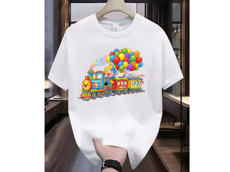K&ouml;nnte beinhalten: Ein wei&szlig;es kurz&auml;rmeliges T-Shirt mit einem bunten Grafikdruck eines Cartoon-Clowns, der eine Lokomotive f&auml;hrt, die zwei Waggons zieht. Der Zug ist mit einem gro&szlig;en B&uuml;ndel bunter Luftballons geschm&uuml;ckt, die &uuml;ber ihm in der Luft schweben.