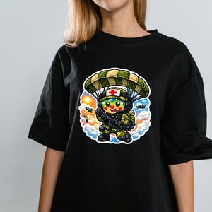 May include: Black t-shirt with a cartoon illustration of a medic soldier parachuting. The soldier wears a helmet with a red cross and holds a weapon. The design includes a parachute, clouds, and a sun. The design is colourful.
