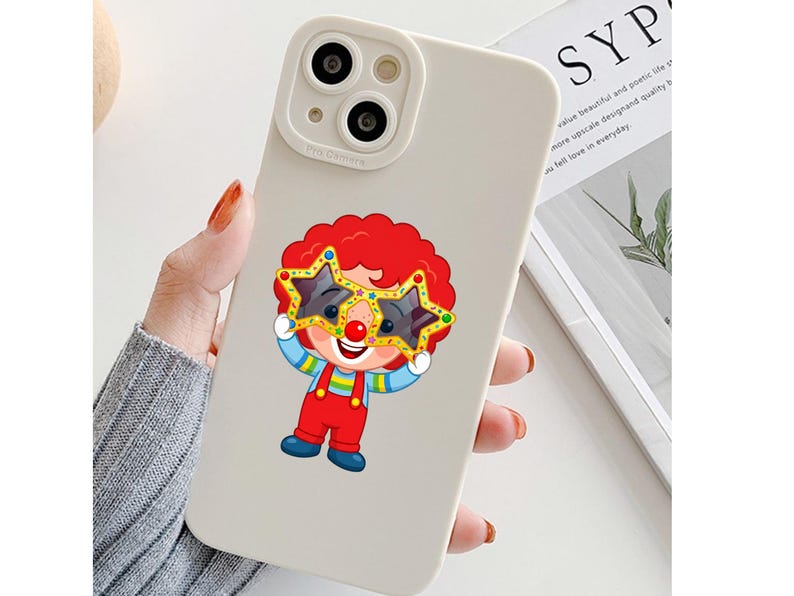 K&ouml;nnte beinhalten: Eine matte, cremewei&szlig;e Smartphone-H&uuml;lle mit einer Cartoon-Clown-Illustration. Der Clown hat leuchtend rotes Haar, eine rote Nase und tr&auml;gt eine gro&szlig;e, gelbe, sternf&ouml;rmige Brille. Er ist mit einem gestreiften Oberteil, einer roten Latzhose und blauen Schuhen bekleidet.