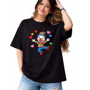 K&ouml;nnte beinhalten: Ein schwarzes T-Shirt mit kurzen &Auml;rmeln und einem Grafikdruck eines l&auml;chelnden Clowns, der einen gestreiften Hut und bunte Kleidung tr&auml;gt, umgeben von mehreren kleinen, mehrfarbigen Herzen in den Farben Rot, Gelb, Blau, Pink und Gr&uuml;n.