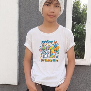 May include: White t-shirt with a colorful graphic featuring a cartoon mouse wearing a party hat, the number 1, and balloons. Text on the shirt reads "Brother of 1st Birthday Boy."