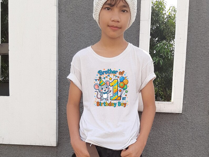 May include: White t-shirt with a colorful graphic featuring a cartoon mouse wearing a party hat, the number 1, and balloons. Text on the shirt reads "Brother of 1st Birthday Boy."