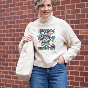 May include: A cream-colored cable knit sweater with a graphic that reads "Grandma of the Birthday Boy" featuring a cartoon mouse holding balloons and the number 1. The person is wearing blue jeans and carrying a cream-colored tote bag.