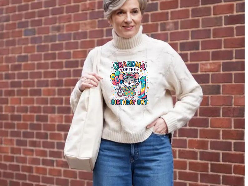 May include: A cream-colored cable knit sweater with a graphic that reads "Grandma of the Birthday Boy" featuring a cartoon mouse holding balloons and the number 1. The person is wearing blue jeans and carrying a cream-colored tote bag.