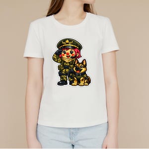 May include: White t-shirt with a cartoon illustration of a soldier saluting, alongside a German Shepherd dog. The soldier is in a green uniform and hat. The dog has a black collar and is sitting. The design is colourful and detailed.