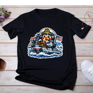 May include: Black t-shirt with a cartoon astronaut captaining a ship. The astronaut wears a white hat and uniform, with an American flag in the background. The ship is grey and red, sailing on blue waves.