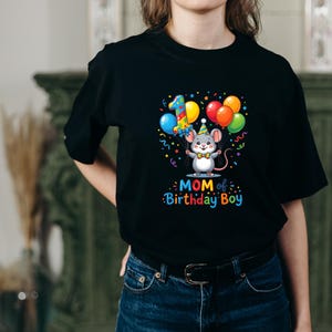 May include: Black t-shirt featuring a colorful graphic of a mouse holding balloons, a party hat, and the number 1. The text "MOM of Birthday Boy" is below the graphic. The shirt is worn by a person.
