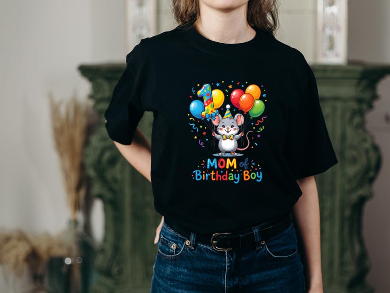 May include: Black t-shirt featuring a colorful graphic of a mouse holding balloons, a party hat, and the number 1. The text "MOM of Birthday Boy" is below the graphic. The shirt is worn by a person.