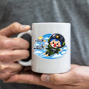 May include: White ceramic mug with a cartoon pilot illustration in a green flight suit, black helmet, and red clown nose. The design includes two fighter jets, clouds, and a sun. The mug is held by a person.