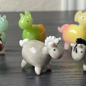 May include: Six small glass figurines of horses in various colors, including brown, gray, green, and yellow. The horses are all standing on a dark surface.