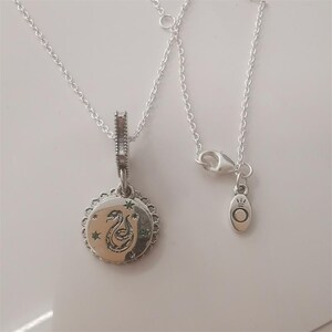 May include: A silver necklace featuring a circular pendant with a snake engraving surrounded by small star motifs. The pendant has a scalloped edge and hangs from a delicate chain. A small oval tag with a crown and circle logo is attached to the clasp.