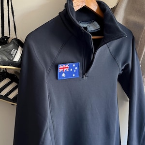 May include: A dark navy blue long-sleeve pullover with a quarter-zip neckline and a high collar. A rectangular patch featuring the blue, white, and red flag of Australia is stitched onto the upper right chest area of the garment.