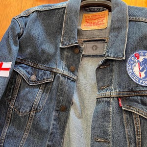 Könnte beinhalten: Blaue Jeansjacke mit einem Chelsea Football Club-Aufnäher und einem England-Flaggen-Aufnäher. Die Jacke hat ein klassisches Design mit Knopfverschlüssen, Taschen und einem Levi Strauss & Co. Label.