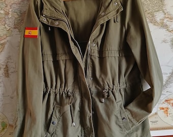 May include: An olive green utility jacket featuring a hood, a front zipper closure with snap buttons, and a drawstring waist. The right sleeve displays a rectangular patch of the Spanish flag. A label is visible at the inner neckline.