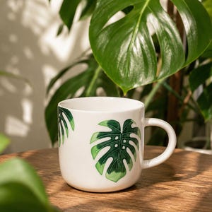 May include: A white ceramic mug featuring a glossy green monstera leaf illustration on the front and side. The mug has a smooth, rounded body and a curved handle, showcasing a botanical design with deep green and light green leaf details.