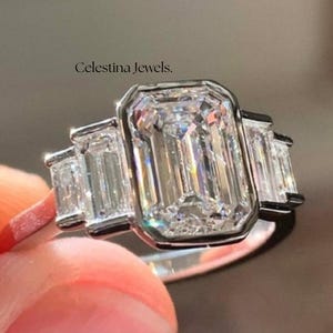 Op de afbeelding: Een platina ring met een smaragdgeslepen middensteen, geflankeerd door twee kleinere baguette-diamanten. De ring is gezet met een halo van kleinere stenen. De tekst "Celestina Jewels" is zichtbaar.