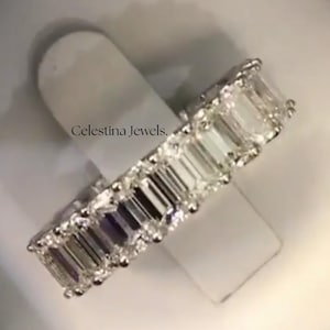 May include: A close-up of a diamond eternity ring with emerald-cut stones. The ring is set in a silver band and is displayed in a red and white jewelry box. The ring is on a white stand.