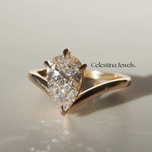 May include: A gold ring with a pear-shaped diamond center stone. The ring has a unique V-shaped band design. The text "Celestina Jewels" is visible on the right side of the image. The ring is set against a light background.
