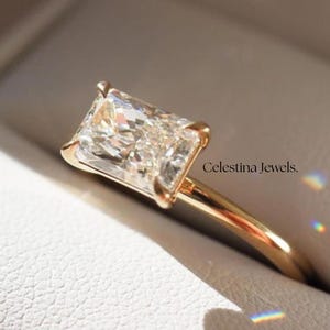 May include: A gold ring with a rectangular-cut diamond is displayed in a white box. The diamond reflects light. The ring is from Celestina Jewels.