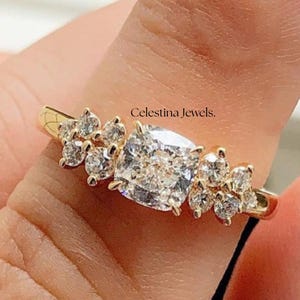 May include: A gold ring featuring a large, square-cut diamond center stone, flanked by clusters of smaller stones. The ring is held up, with the text "Celestina Jewels" visible.