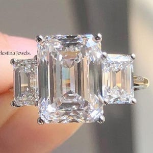 Può includere: Un anello di fidanzamento a tre pietre con una pietra centrale taglio smeraldo affiancata da due pietre laterali più piccole taglio smeraldo. L'anello ha una fascia d'oro ed è incastonato con gemme chiare e scintillanti. Il testo "Celestina Jewels" è visibile.