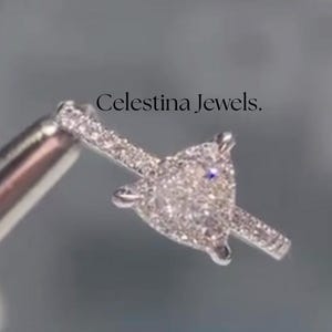 May include: A diamond ring with a heart-shaped central stone, held by a silver tool. The ring is encrusted with small, sparkling diamonds. The text "Celestina Jewels" is visible in the background.