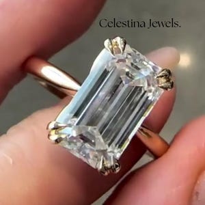 May include: An emerald cut diamond ring with a gold band. The clear, rectangular diamond is the focal point, set in a gold band. The text "Celestina Jewels" is visible in the image.