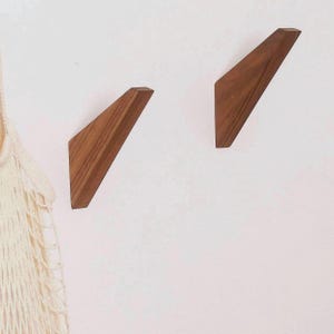 May include: Two angled wall hooks made of brown wood with a visible grain pattern. The hooks have a triangular shape with a flat base and a slanted top edge, mounted separately on a plain surface next to a cream-colored mesh fabric.