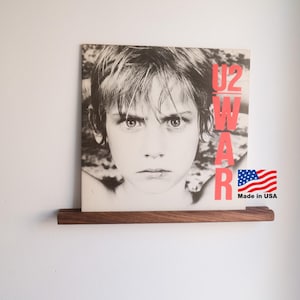 May include: A framed album cover for U2's "War" album, featuring a black and white portrait of a young person with the word "WAR" in red. The album is displayed on a dark wood shelf. A small "Made in USA" sticker with the American flag is visible.